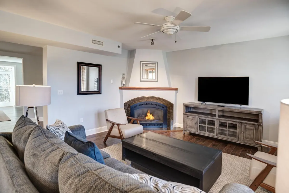The image features a warm, inviting living room with a crackling fireplace, a plush sofa, stylish coffee table, and a TV, all arranged for comfort and socializing. The setting is perfect for relaxing evenings, game nights, or movie marathons after exploring Asheville or the nearby Biltmore Estate.