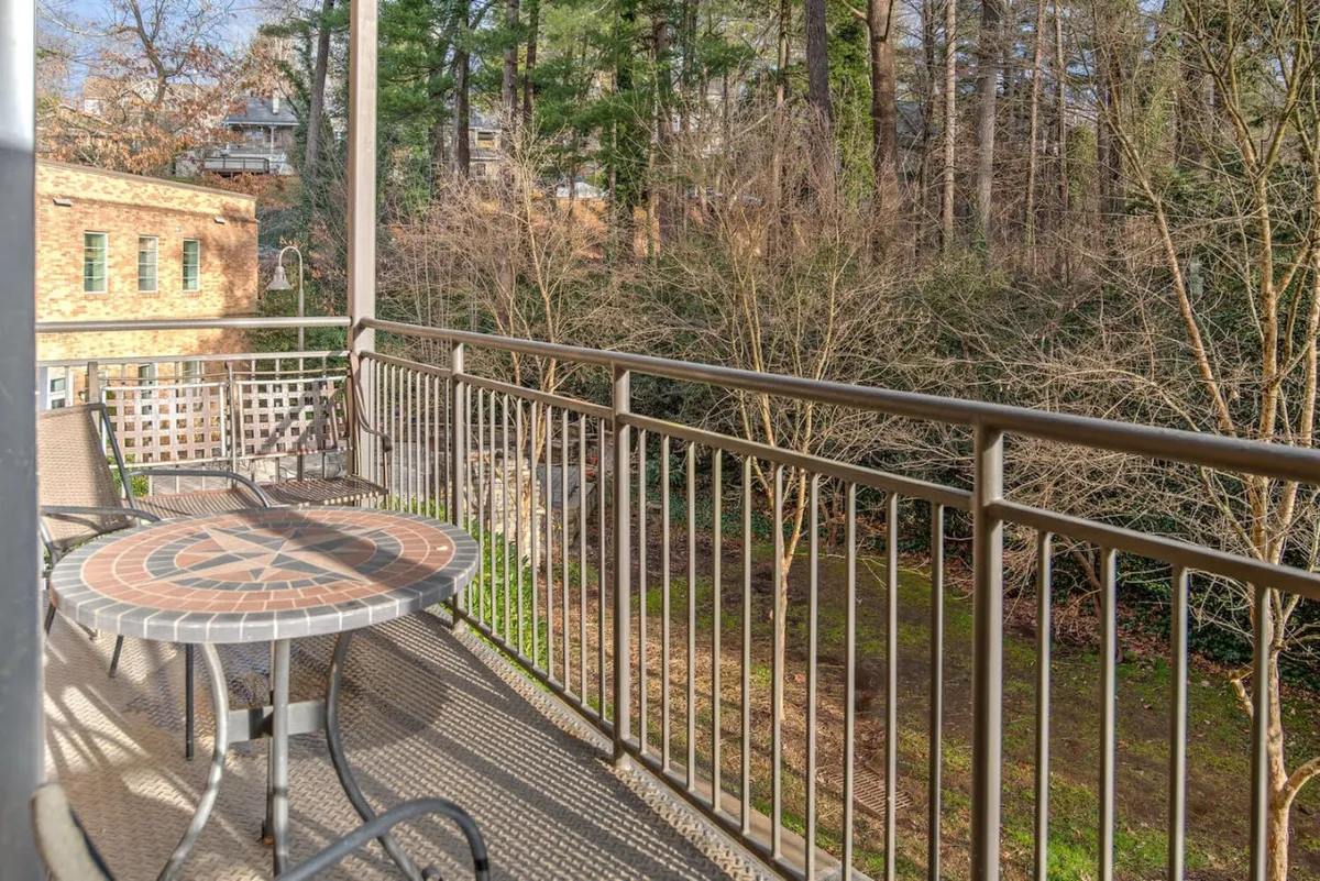 This image features an exterior table with a mosaic top set beside a beautiful metal railing, surrounded by lush greenery. The scene feels welcoming and offers a tranquil space for relaxation or conversation, with natural light illuminating the area.
