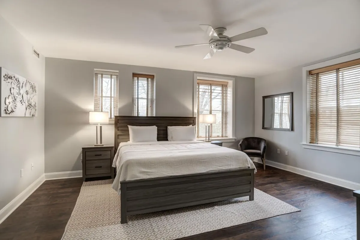A tranquil bedroom with a king-sized bed, nightstand, ample natural light streaming through large windows, a chair, and a mirror. The room radiates peace and offers a cozy spot for relaxation or quiet conversation.
