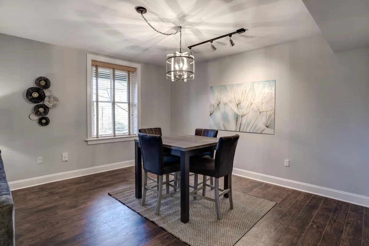 This image presents a cozy dining area with a prominent dining table, a chandelier overhead, wall artwork, and a window with blinds filtering in gentle light. The setting is designed for family meals, social gatherings, and celebration, offering a welcoming ambiance that complements the adjacent kitchen space.