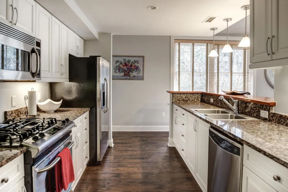 This image displays a bright, well-equipped kitchen featuring stainless steel appliances, granite countertops, a kitchen island with seating, and a window allowing natural light to flow in. Framed artwork and neatly organized elements create a welcoming and functional atmosphere. The visible kitchen island suggests ample space for meal prep and casual dining, tying together modern convenience with a warm, homey ambiance.