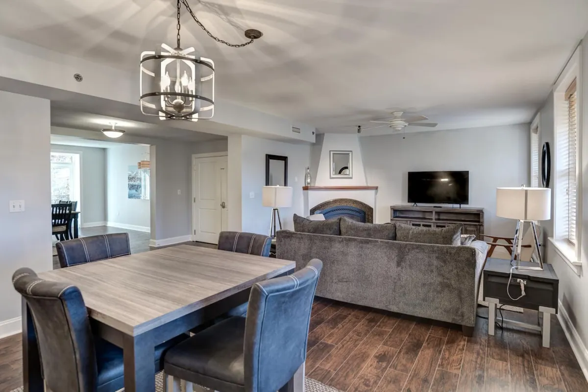 This image shows a spacious, open-plan area combining a dining table, sofa, fireplace, and television. Large windows bring in natural light, accentuating the blend of original details and modern updates. The layout encourages both socializing and relaxation, ideal for friends and families to gather after a day exploring Asheville.
